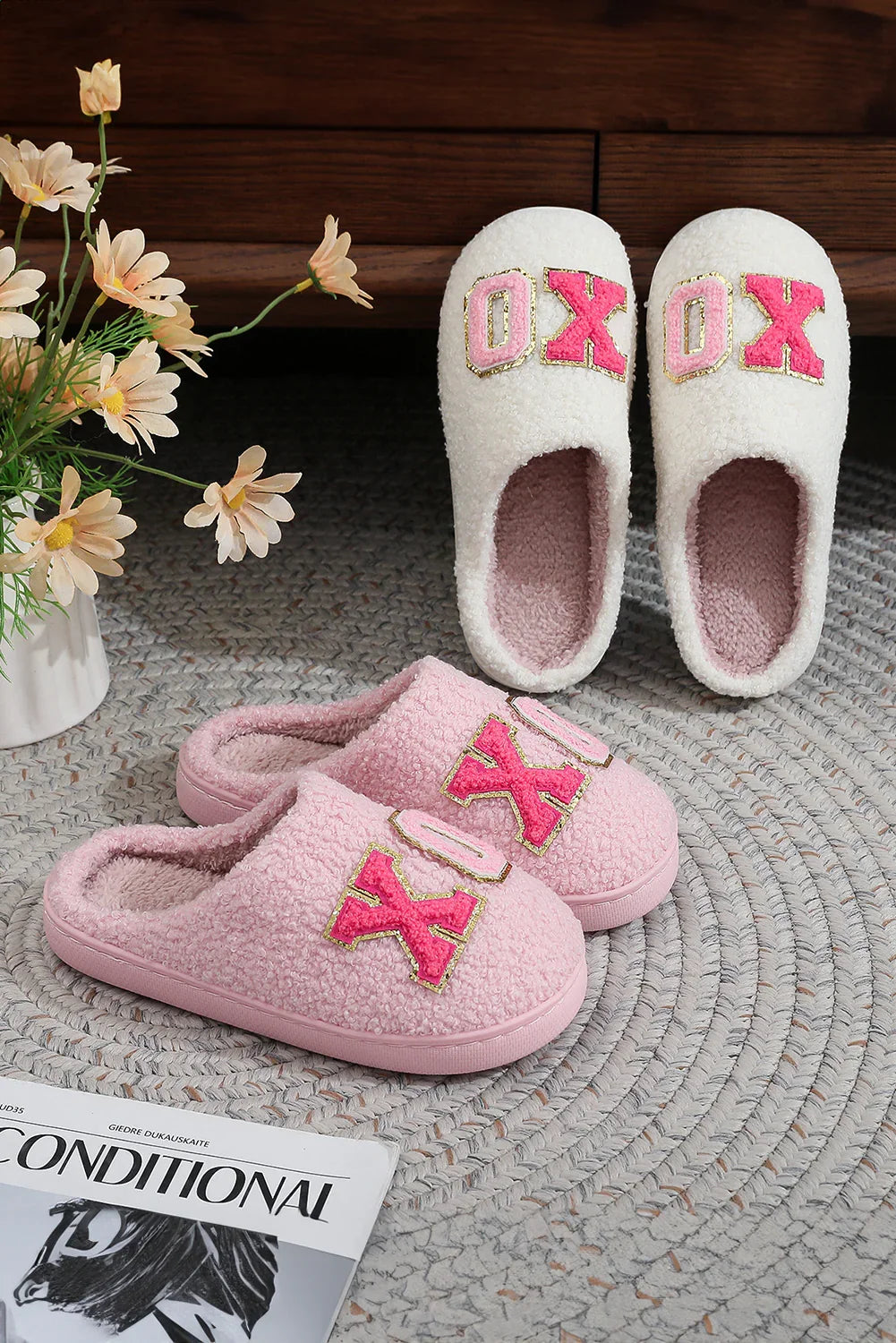 Cozy pink and white fleece slippers with XOXO design on a braided rug near flowers and magazine