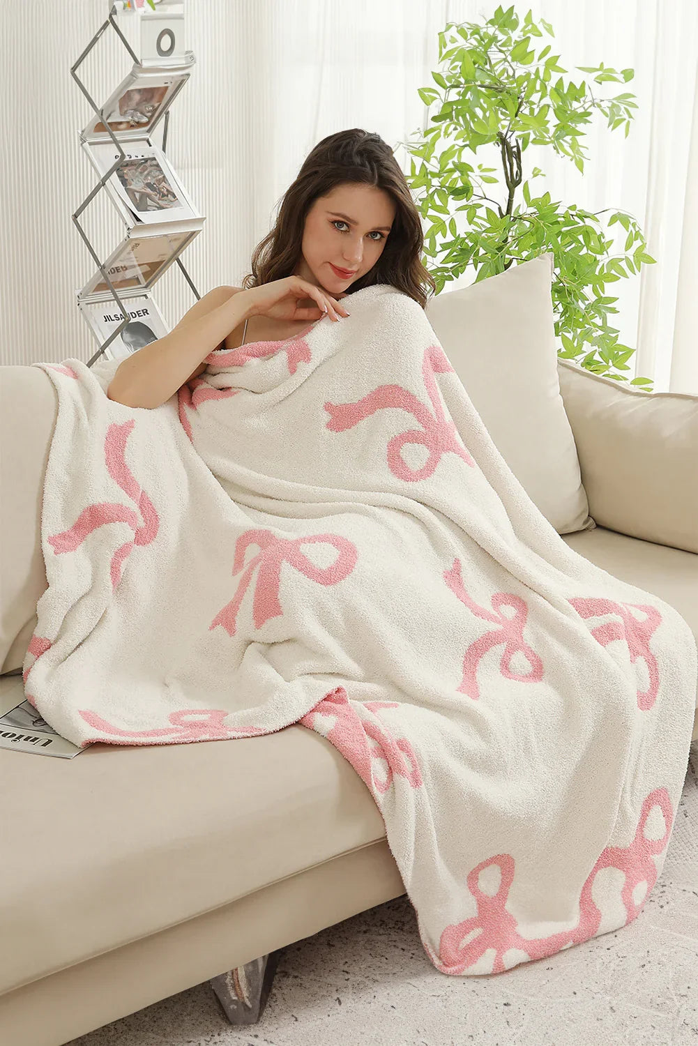 Woman wrapped in soft white blanket with pink bows sitting on beige sofa in bright living room with green plant