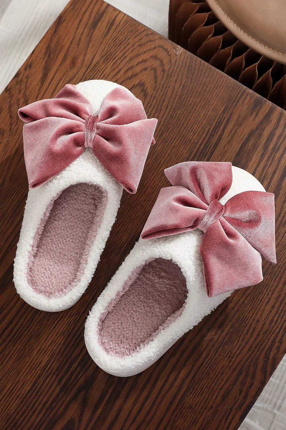 Pair of white fuzzy slippers with large pink velvet bows on a wooden surface
