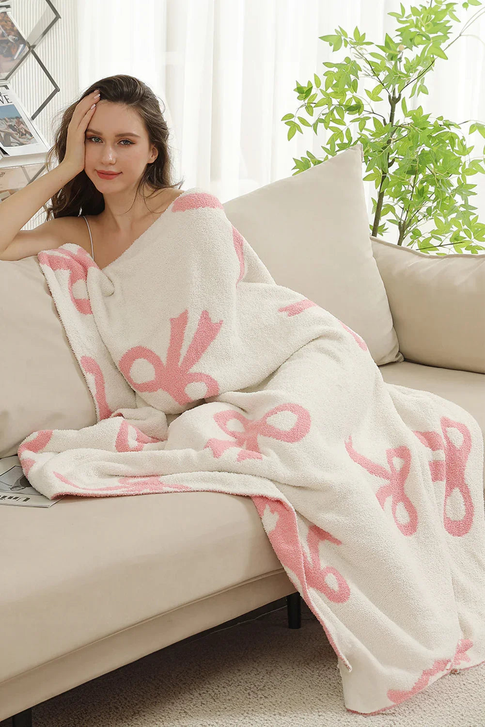 Young woman wrapped in cozy white blanket with pink bow patterns lounging on beige sofa