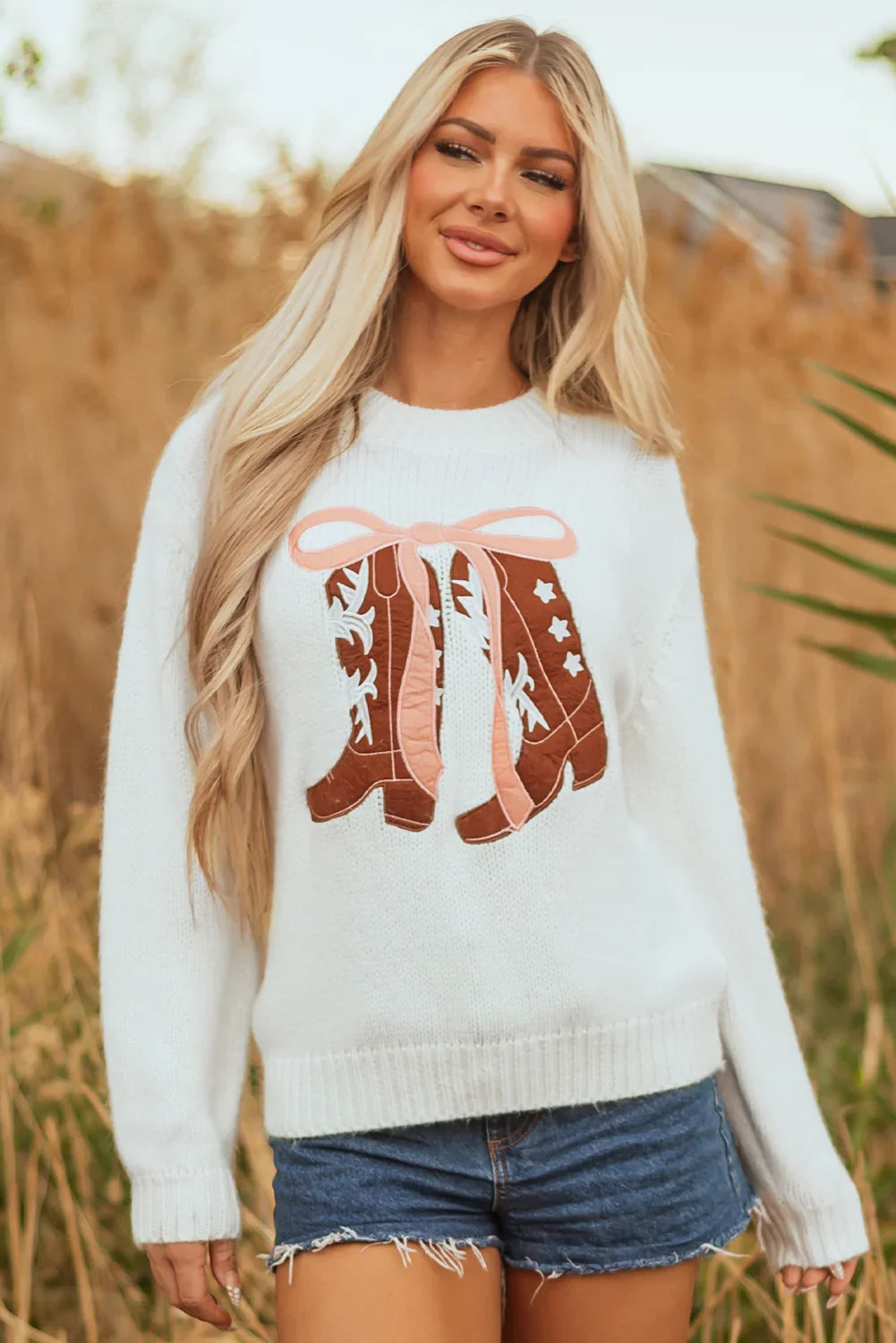 Blonde woman wearing white sweater with brown cowboy boots design and denim shorts outdoors