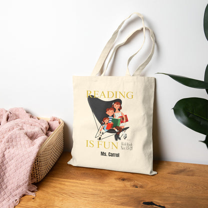 A cream canvas tote bag with a vintage-style illustration of a woman reading to children in a stroller, with the text 'READING IS FUN' and 'Ms. Carrol'.
