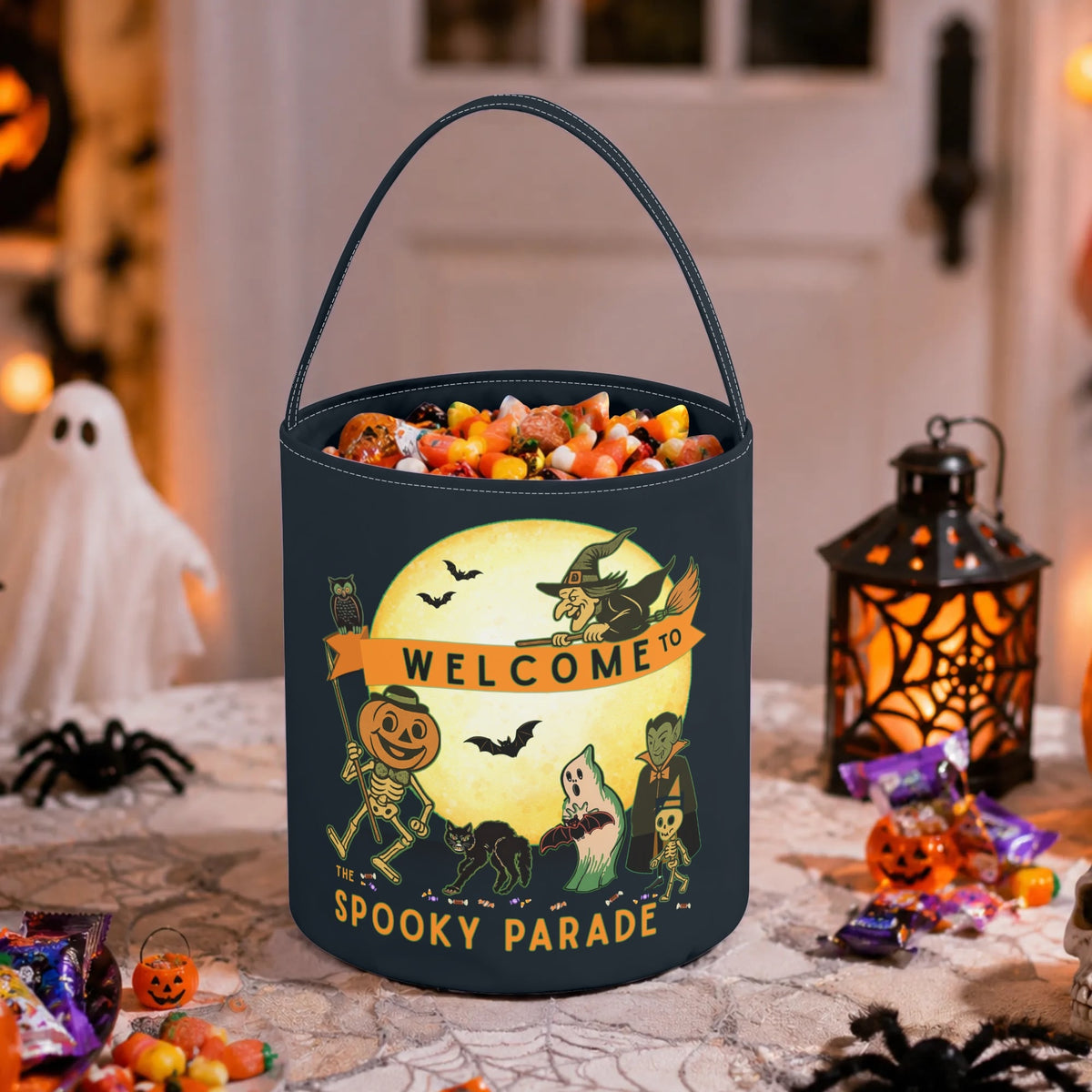 A black cylindrical Halloween candy bucket filled with assorted treats, placed on a decorated table. The bucket features a spooky graphic with a yellow moon, a witch, a skeleton, a ghost, and the text 'WELCOME TO Spooky Parade'.