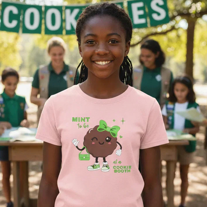 Girl wearing a pink t-shirt with a cookie design, standing in front of a Girl Scout cookie sale event.