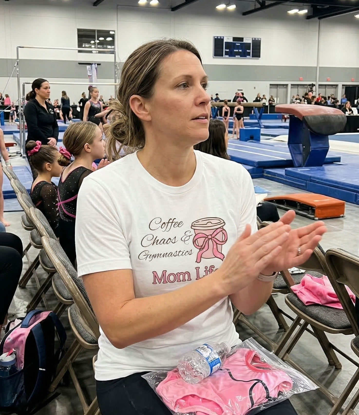 Woman clapping at a gymnastics event with equipment and spectators in the background