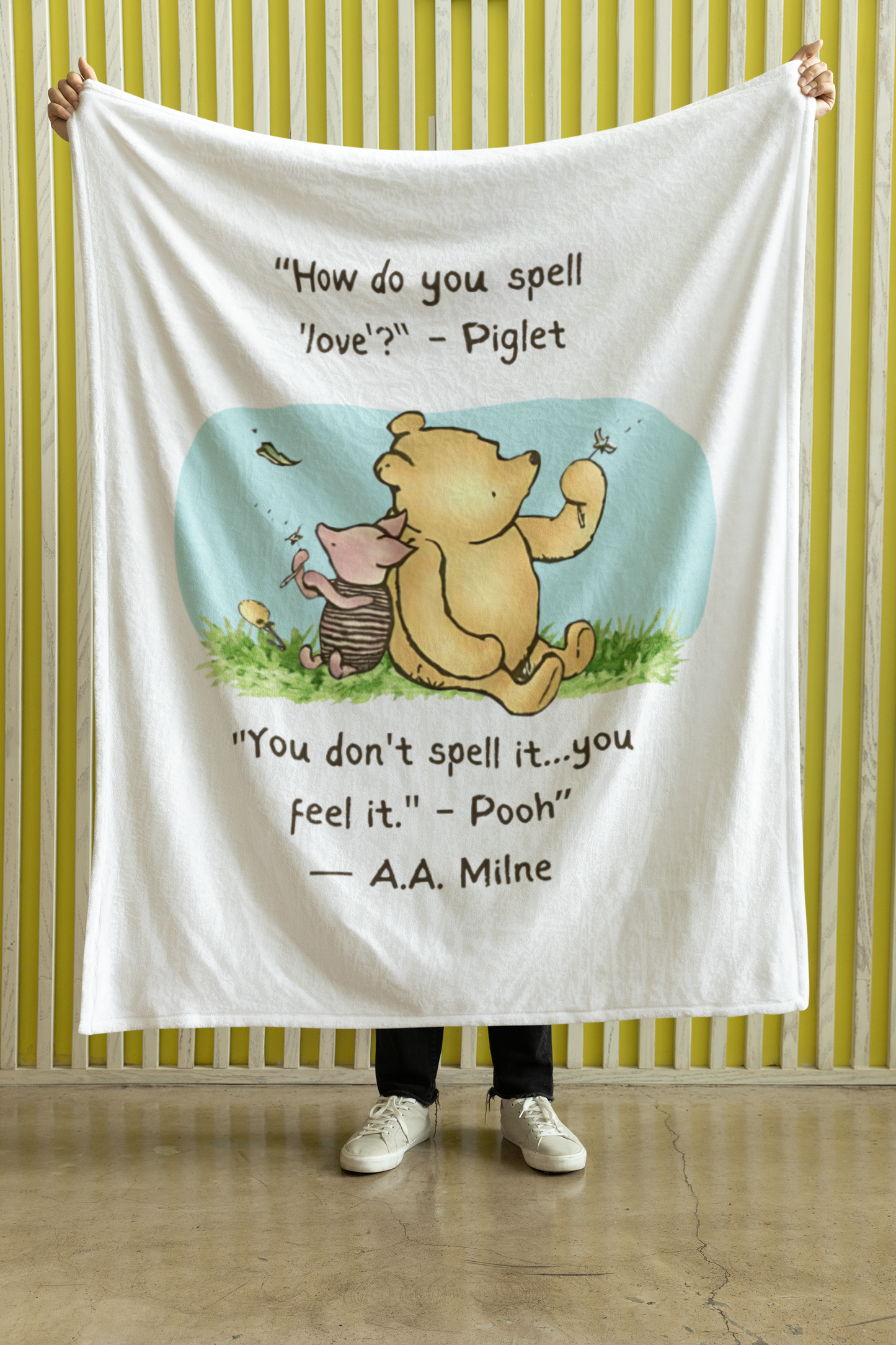 Person holding a Winnie the Pooh and Piglet 50" x 60" blanket with a quote against a striped wall.