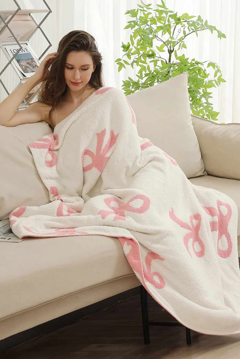 Woman wrapped in white blanket with pink bow patterns sitting on beige sofa in cozy living room