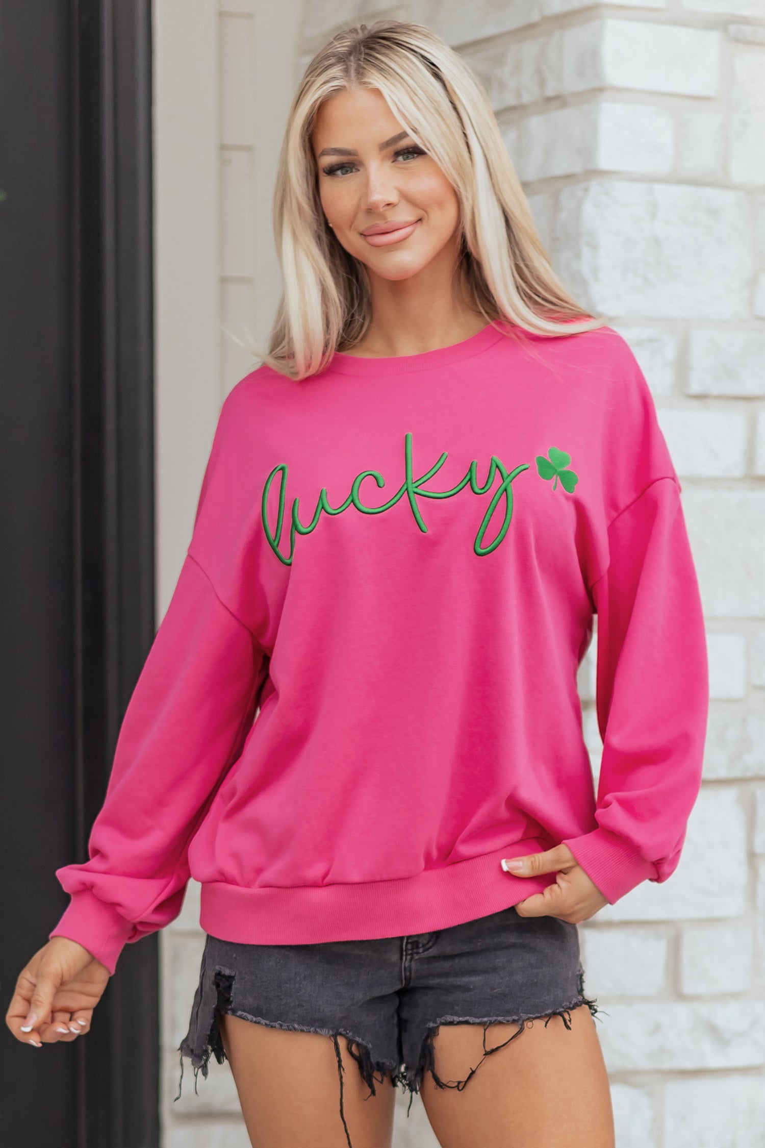 Woman wearing a pink sweatshirt with 'lucky' text, standing against a brick wall.