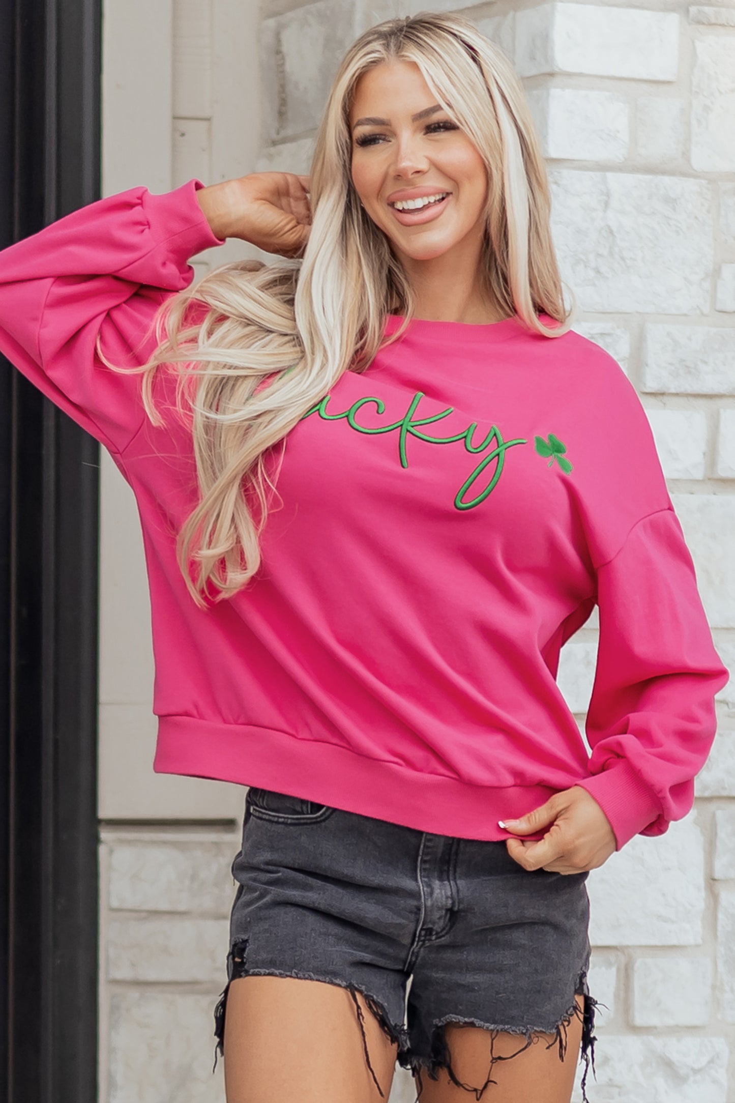 Woman wearing a pink sweatshirt with 'lucky' printed on it, standing against a brick wall.
