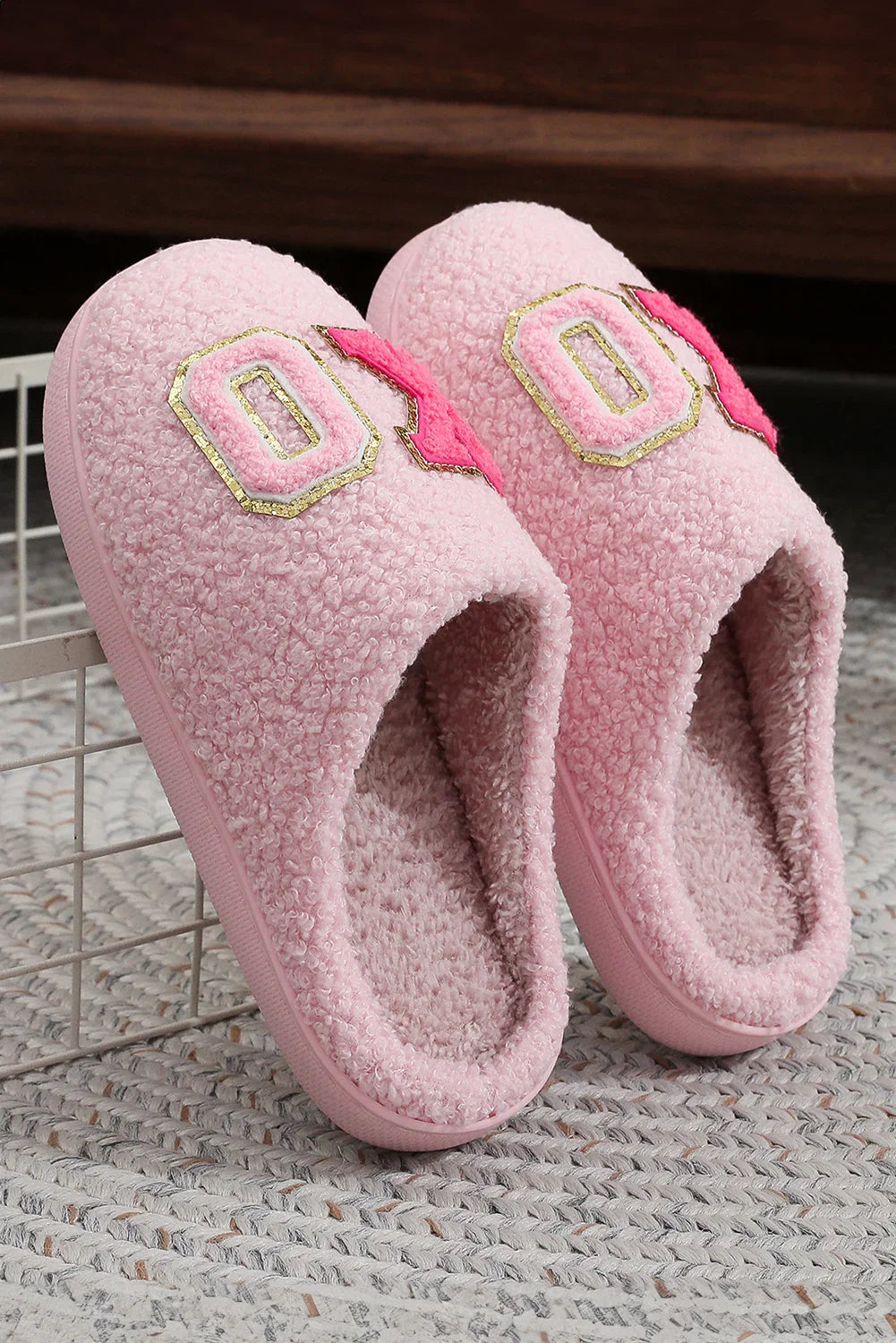 Pair of pink fuzzy indoor slippers with textured exterior and decorative patches, resting on woven rug