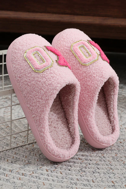 Pair of pink fuzzy indoor slippers with textured exterior and decorative patches, resting on woven rug