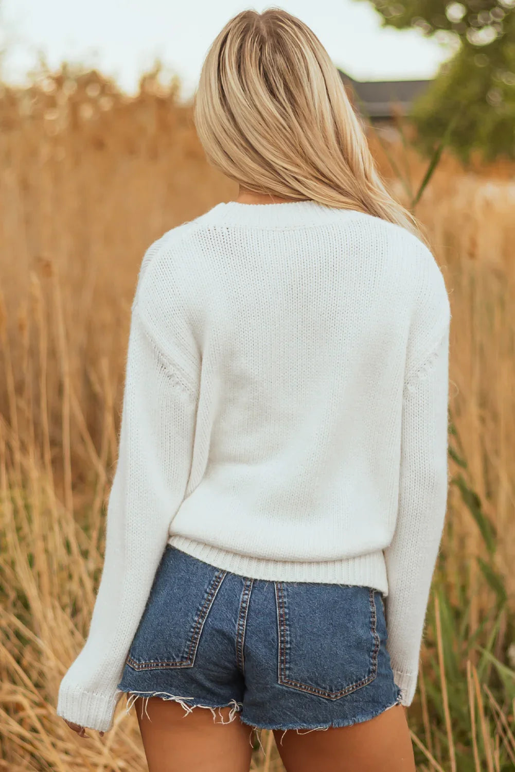 Woman with blonde hair wearing a white knit sweater and denim shorts in a natural outdoor setting