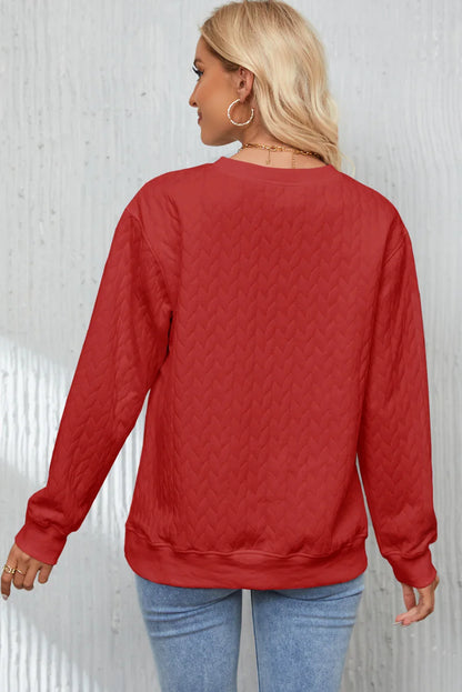 Blonde woman wearing red textured long sleeve sweatshirt and light blue jeans against gray wall