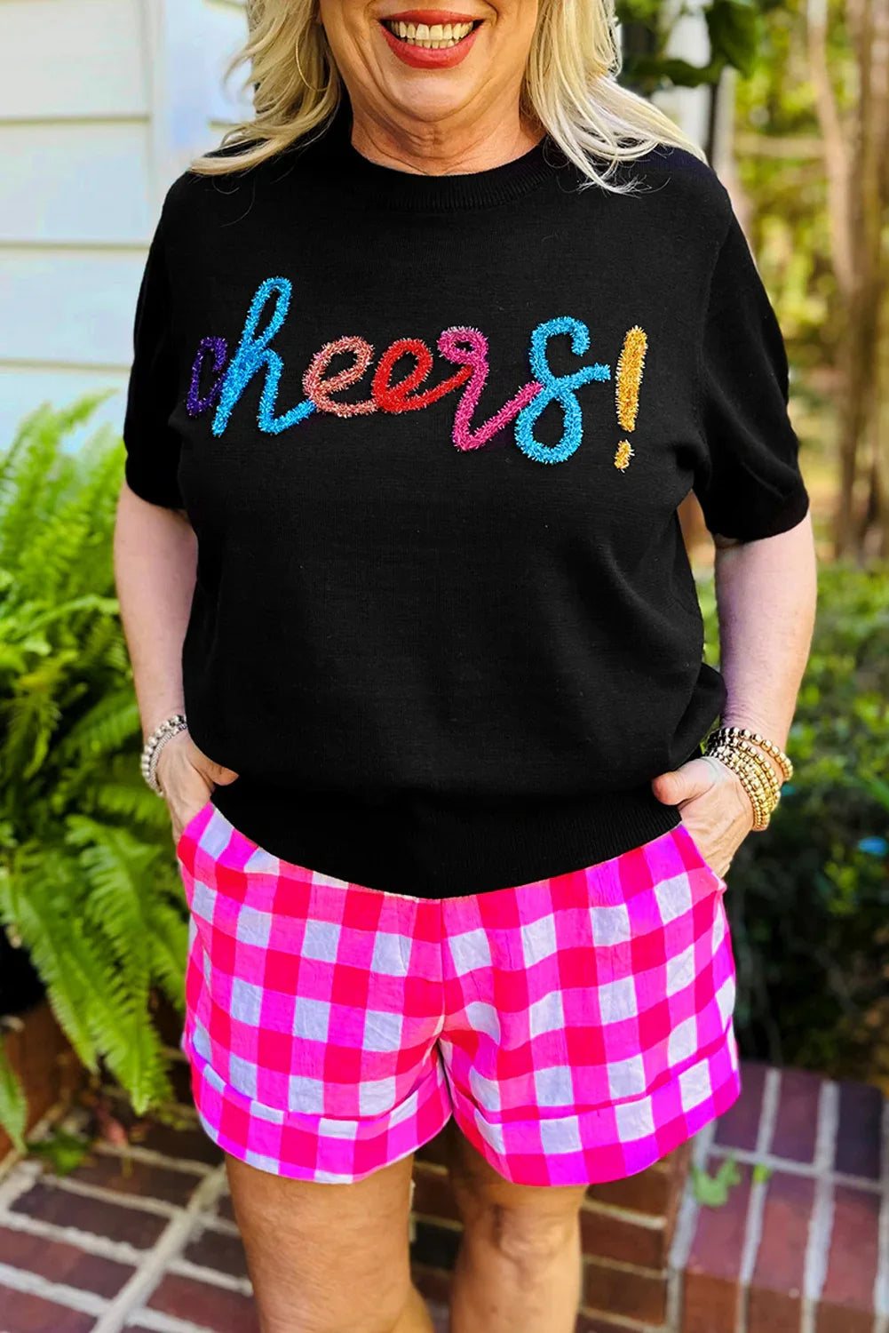 Woman wearing black sweater with multicolor Cheers! text and pink checkered shorts outdoors