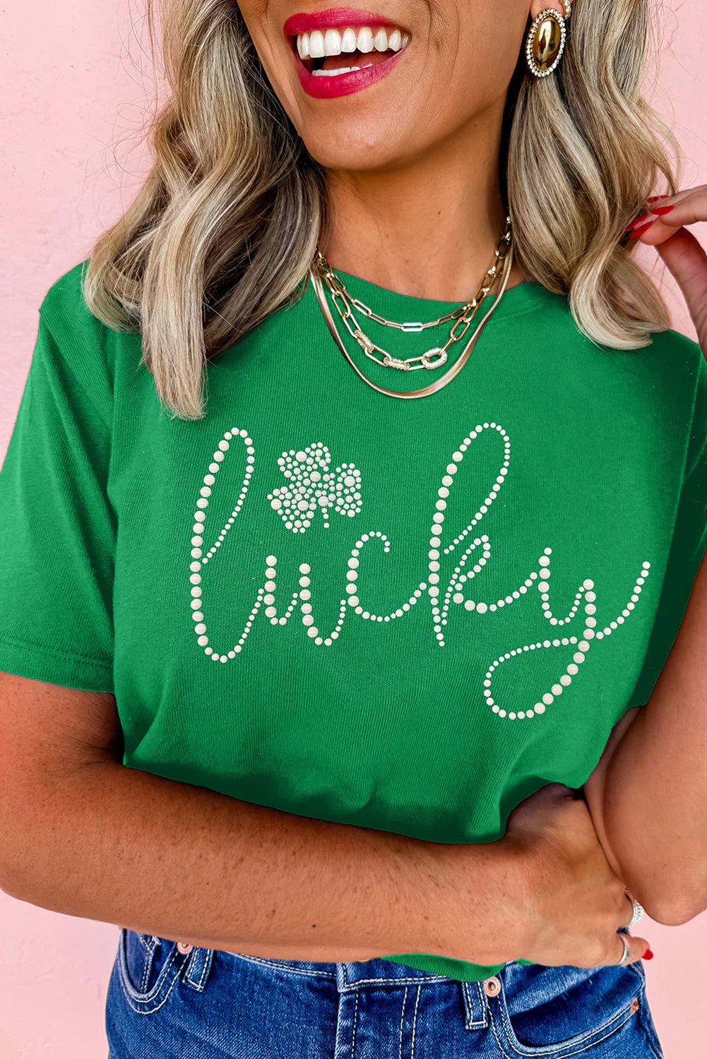 Smiling woman wearing green Lucky t-shirt with pearl accents, gold necklaces, and denim jeans against pink wall