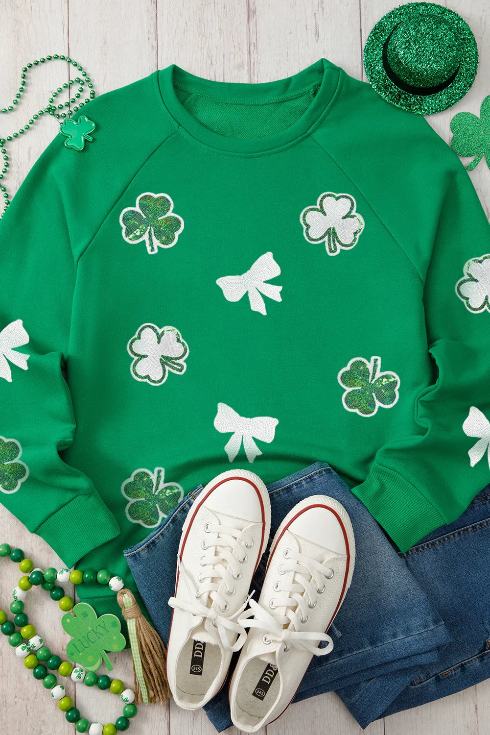 Green sweatshirt with shamrock and bow patterns, paired with white sneakers and St. Patrick's Day accessories on a wooden surface.