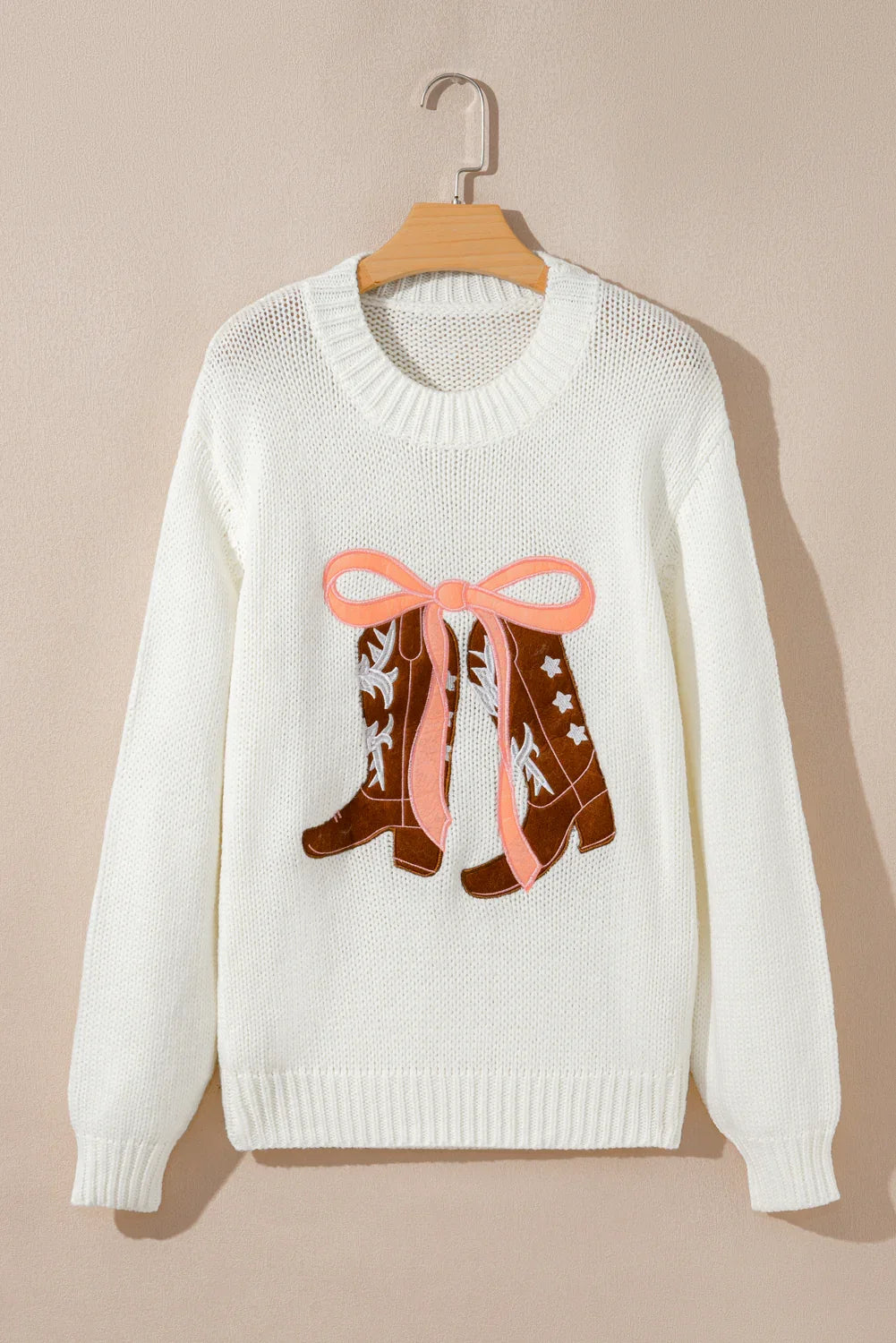 White knitted sweater with embroidered brown cowboy boots and pink bow hanging on wooden hanger