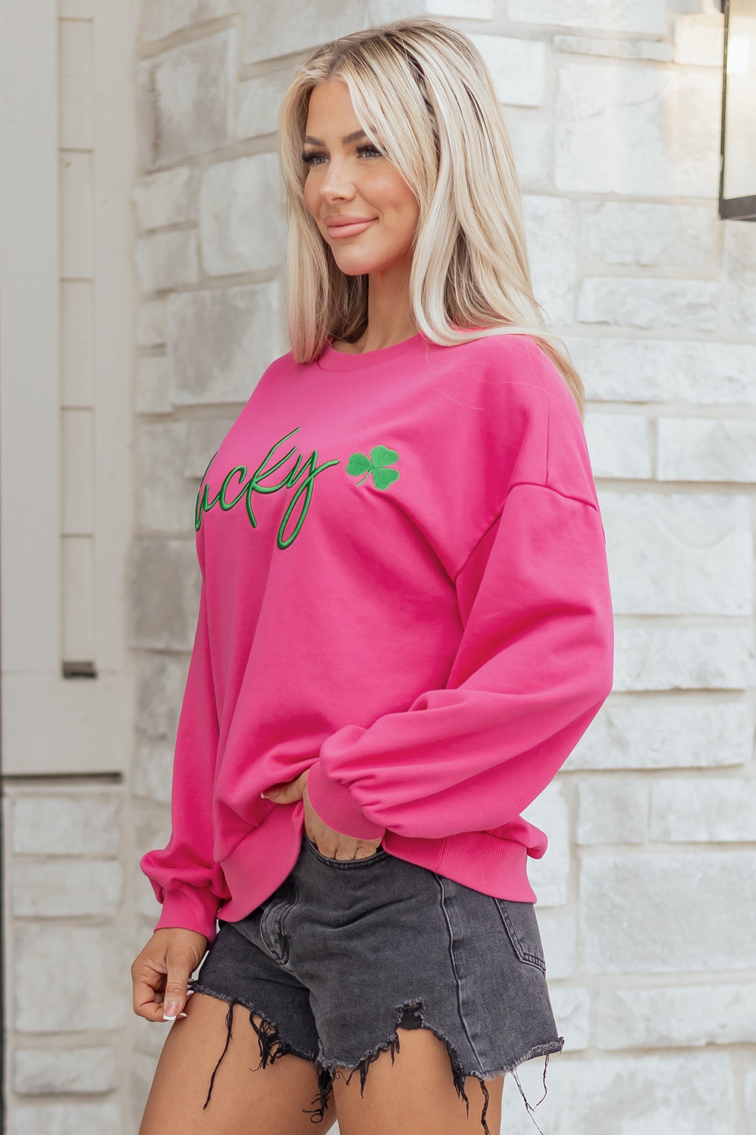 Woman wearing a pink sweatshirt with text and a green clover graphic against a stone wall.