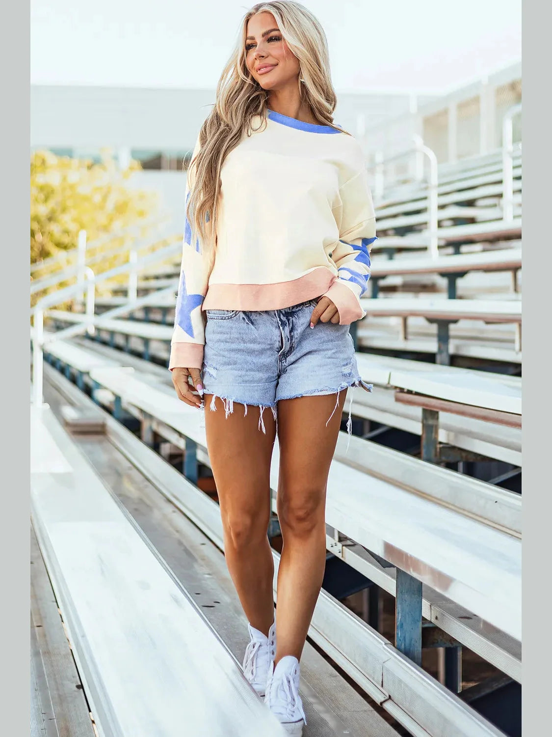 Woman in a colorful sweater and denim shorts standing on bleachers.