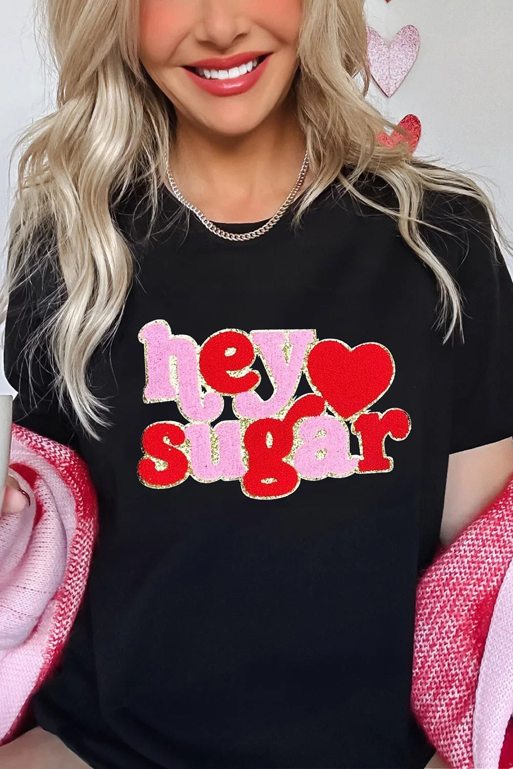 Blonde woman smiling wearing black graphic tee with pink and red text hey sugar and holding pink red knit blanket