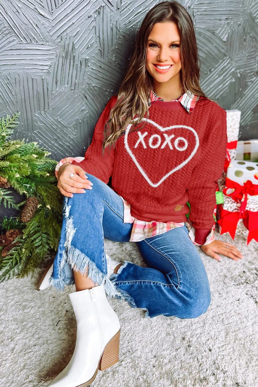 Smiling woman wearing red XOXO sweater and plaid shirt with fringed jeans and white boots, festive gifts and greenery backdrop
