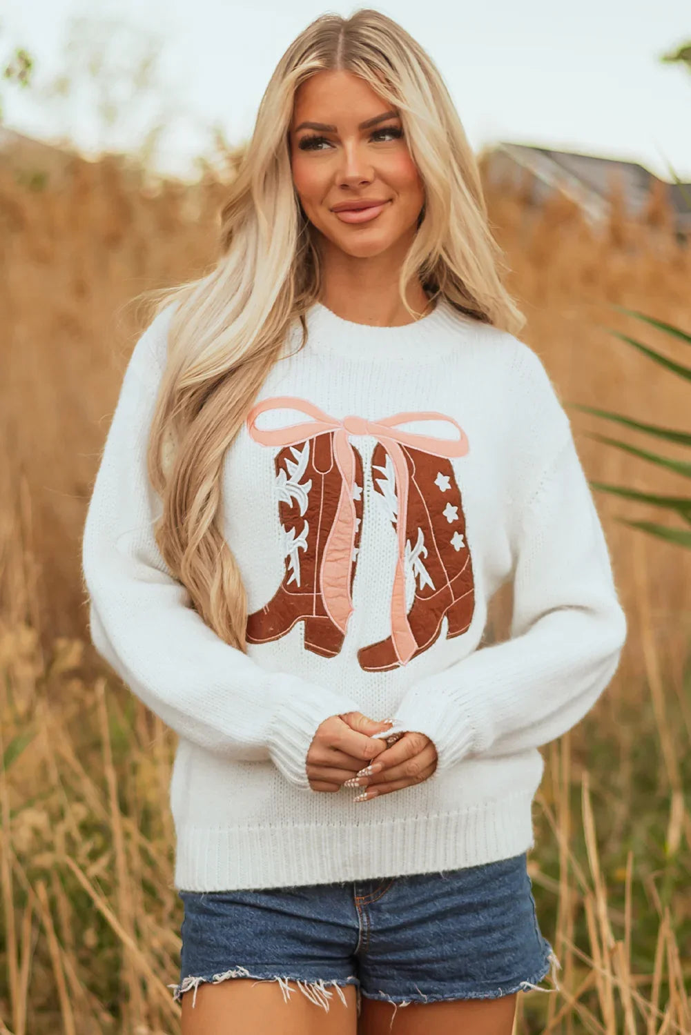 Blonde woman outdoors wearing white sweater with brown cowboy boots design and denim shorts