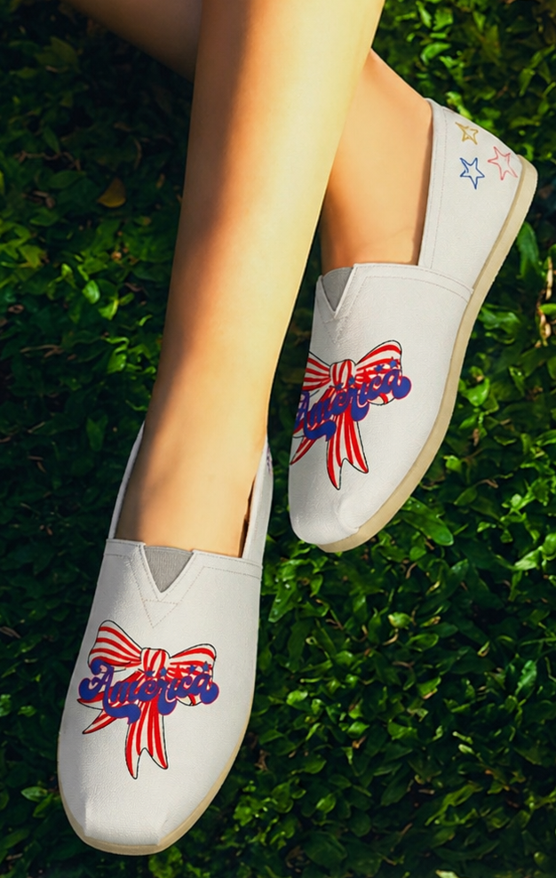 Person wearing white shoes with red, white, and blue America text and red and white strip bow designs a Bow design sitting on grass - close up