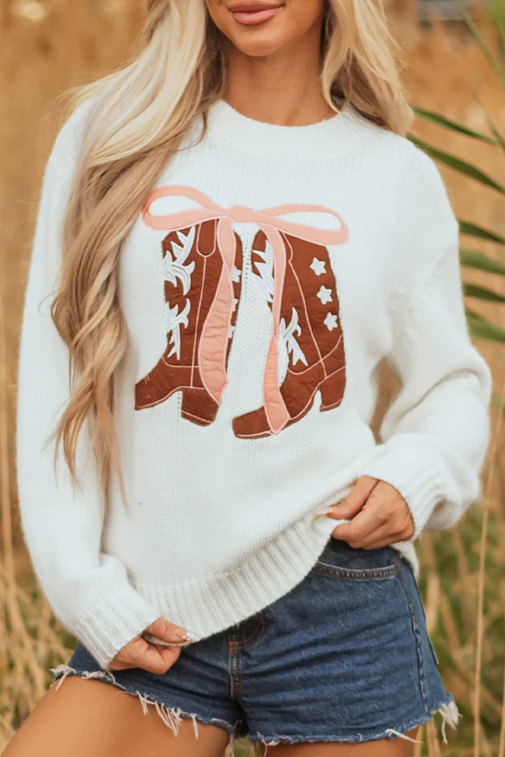 Woman wearing white sweater with brown and pink cowboy boot design paired with denim cut-off shorts