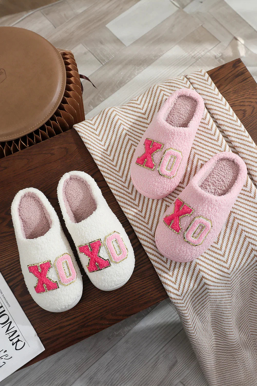 Pair of cozy pink and white fuzzy slippers with XO letters on wooden furniture and chevron blanket