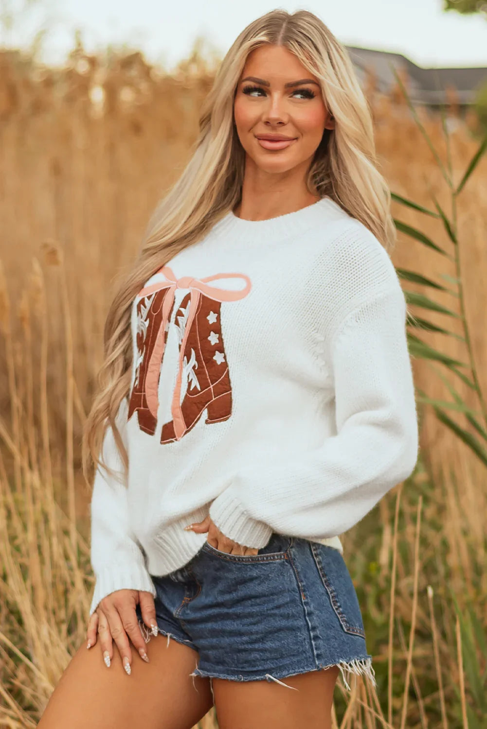 Blonde woman wearing white sweater with western boot design and denim shorts in outdoor field