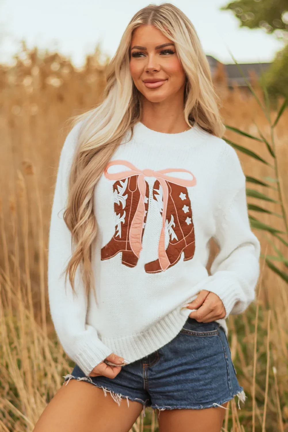 Blonde woman wearing white sweater with cowboy boots design and denim shorts in a field