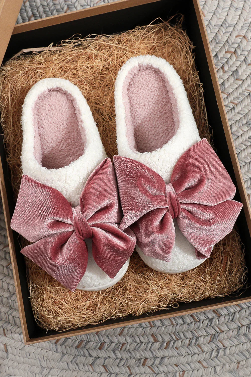 White cozy slippers with large pink velvet bows, displayed in a gift box with straw filling