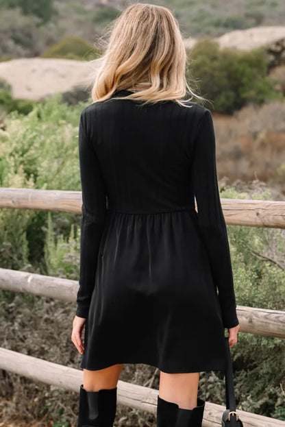 Woman with blonde hair wearing a black long-sleeve dress and black boots outdoors by wooden fence