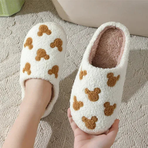 Cozy white fleece slippers with brown teddy bear patterns on a textured carpet