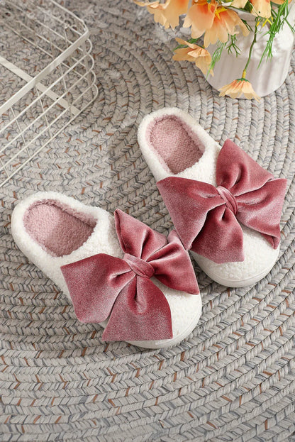Cozy white fuzzy slippers with large pink velvet bows on a braided gray rug near a flower vase