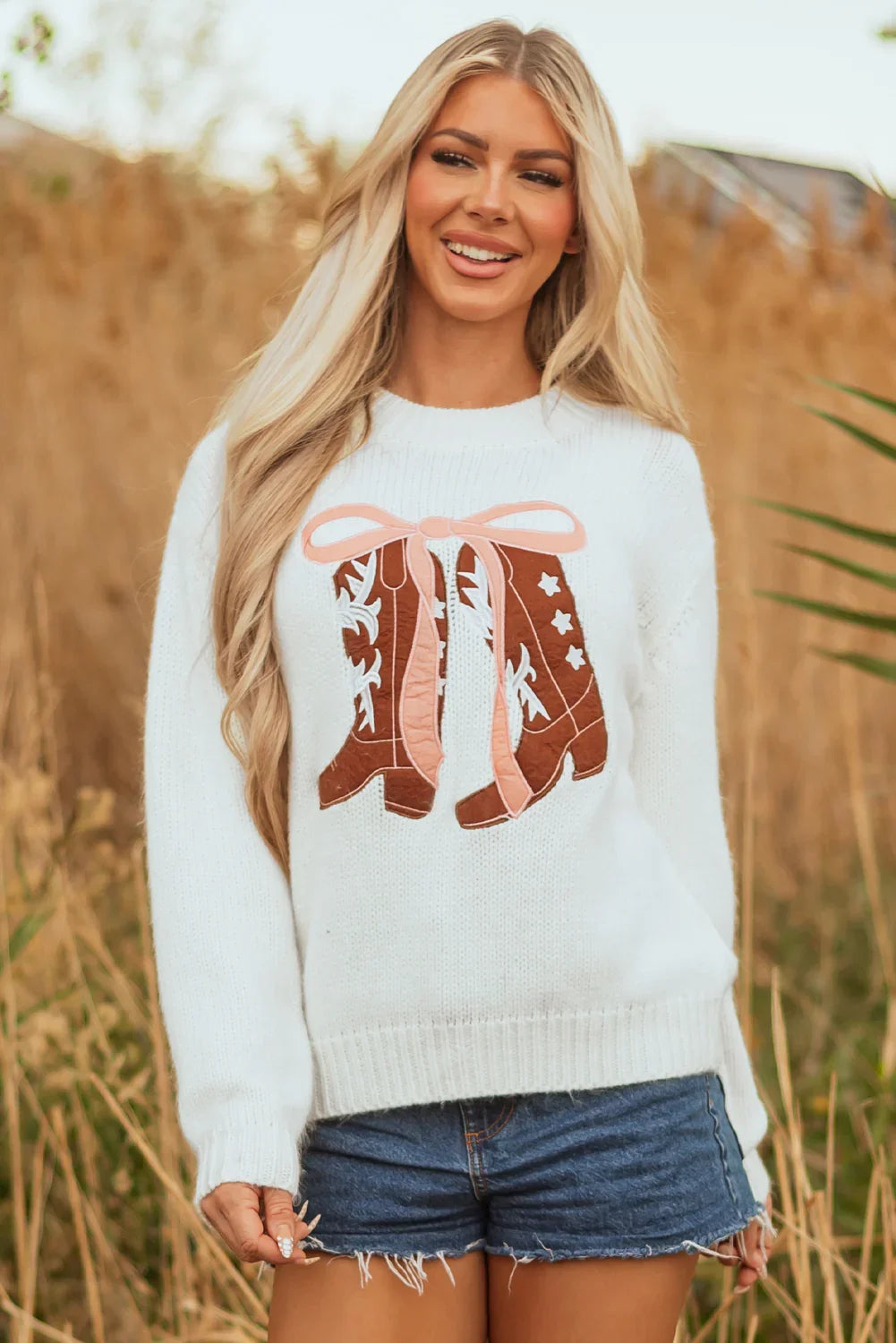 Blonde woman wearing white sweater with cowboy boots graphic and denim shorts outdoors