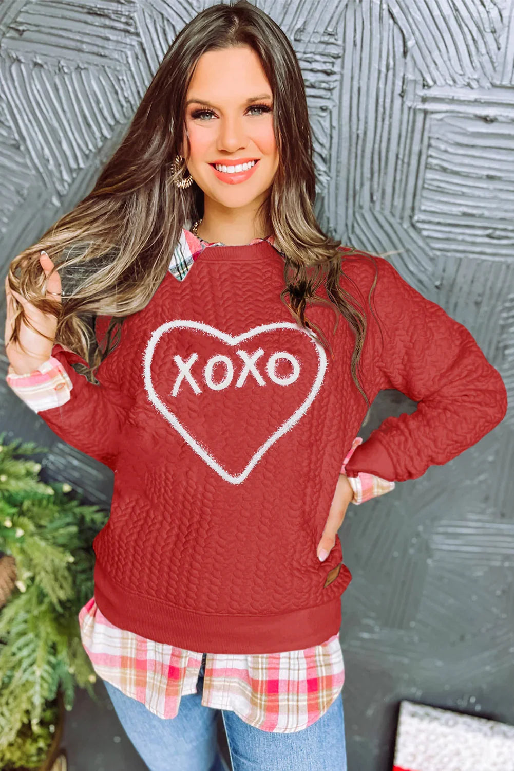 Smiling woman wearing red XOXO sweater over pink plaid shirt with gray textured wall background
